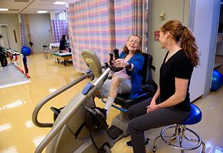 Physical therapist observes as an older woman uses a stationary bike in rehabilitation gym