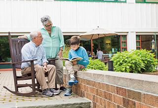 Grandparents spend time reading with a grandchild in the courtyard of a Hebrew SeniorLife senior living community.