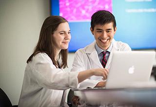 A female and a male Harvard Medical School fellow sit at a table at Hebrew SeniorLife looking at a laptop. They are wearing white coats.