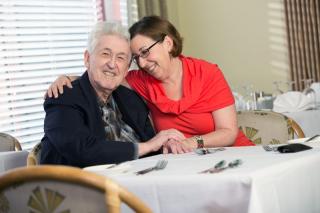 A male senior living resident and his adult daughter embrace in a hug at a dining table.