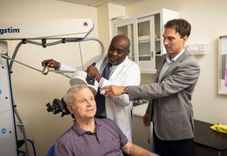 An elderly patient receives TMS treatment for depression at the Wolk Center for Memory Health in Boston. He is seated with a calm expression on his face. A technician holds an electromagnetic coil next to his head while the neuropsychologist looks on.