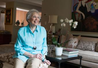 A smiling Orchard Cove resident in a turquoise shirt sits on a couch in her well-appointed independent living apartment.