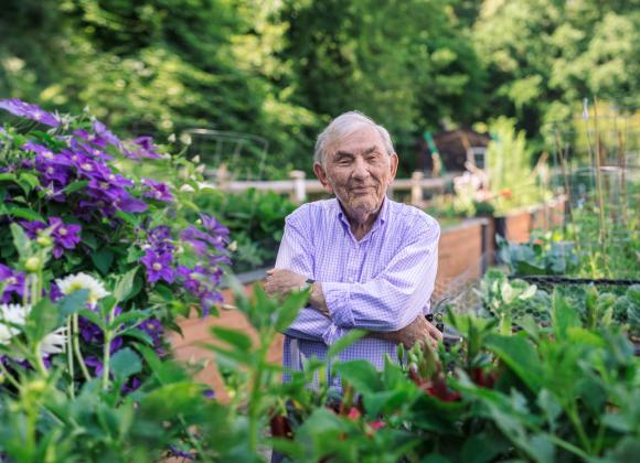 A man standing in the middle of a community garden at NewBridge on the Charles. He is looking at the camera, arms crossed, with a slight smile.