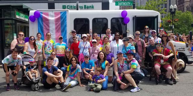 A group of about 30 Hebrew SeniorLife staff and residents wearing colorful outfits standing in front of a Hebrew SeniorLife van draped with a blue-, pink-, and white-striped transgender pride flag at Boston Pride.