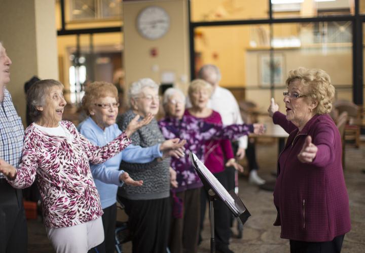 A resident-led chorus practices singing in a brightly-lit community room.