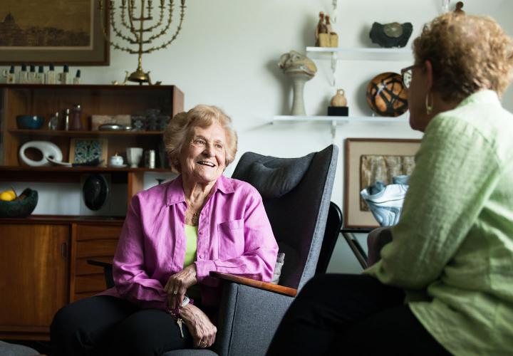 An older woman in a purple shirt sits on a couch in her apartment having a conversation with a friend.