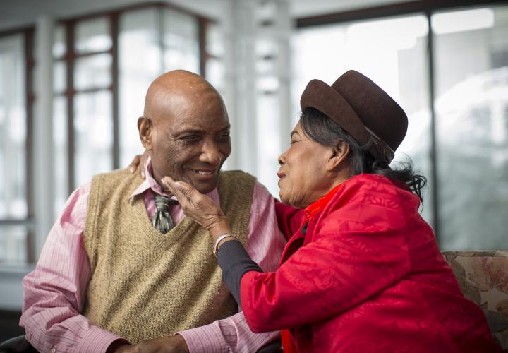 An older man and woman sit in the lobby at Hebrew Rehabilitation Center in Boston; they are smiling at each other, with the woman having her arm around the man and her hand on his face