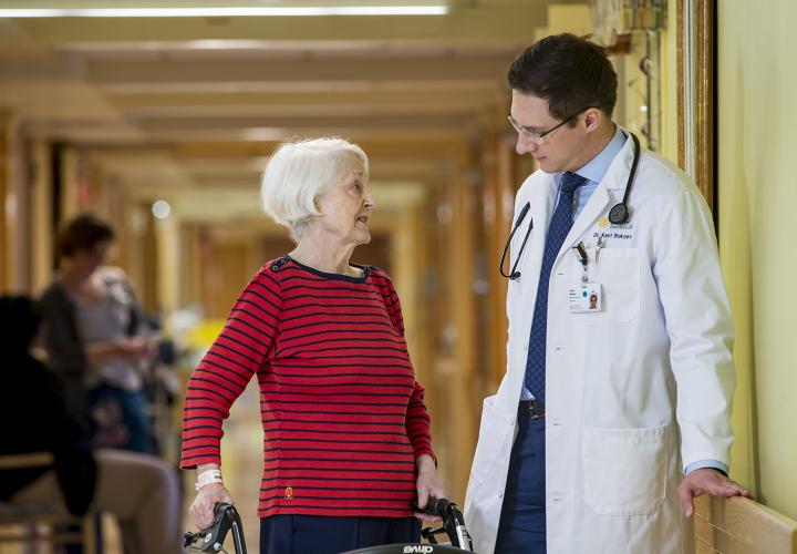 A doctor wearing a white coat leans against the wall in a hallway, talking to an older patient standing next to him.