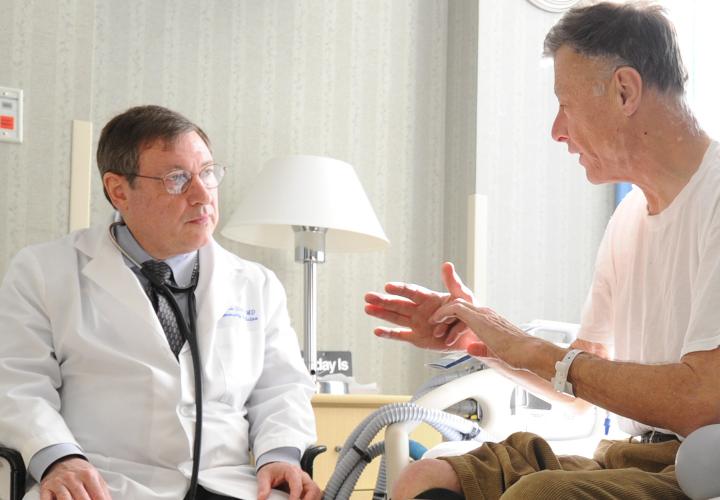 Doctor in white coat sits with patient in sunny hospital room as patient shares his care goals.