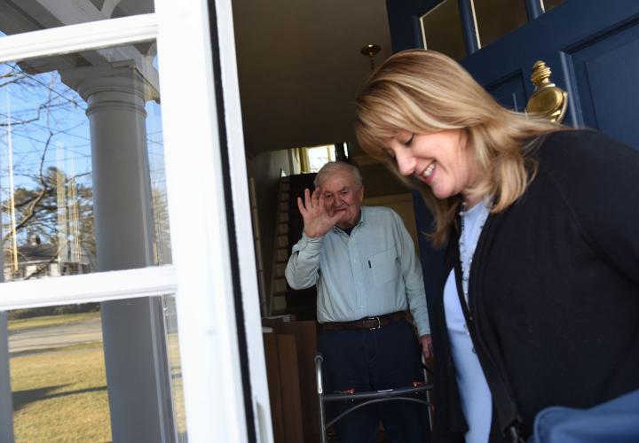 Older man with walker stands at doorway of his home as he waves goodbye to his home health nurse