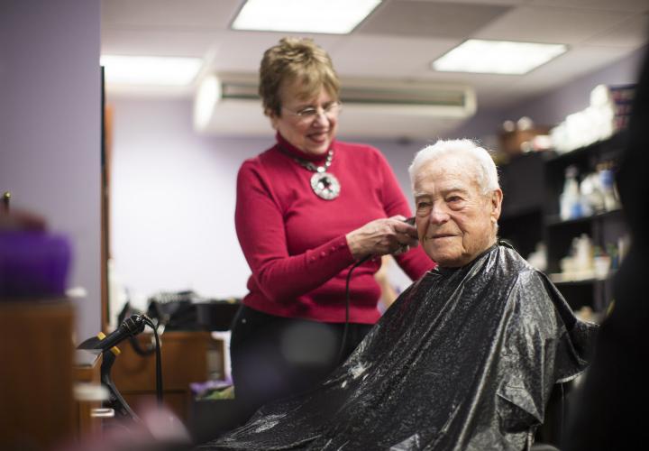 An older man is seated in a barber’s chair wearing a cape. A woman stands behind him, cutting his hair.