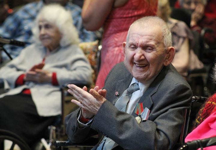 An older Russian man sits clapping and smiling at Hebrew Rehabilitation Center during a Victory Day celebration. He is wearing medals that show he is a veteran.