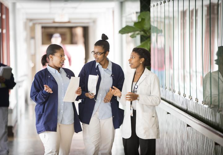 Two female student nurses and a female nurse stand talking in a hallway at Hebrew Rehabilitation Center in Boston. The students are wearing short blue coats and the nurse is wearing a white coat.