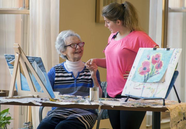 An older woman sits at a table painting at Hebrew Rehabilitation Center in Dedham, with bright artwork in the foreground. She is smiling up at a younger female staff member who is standing next to her and holding her hand.