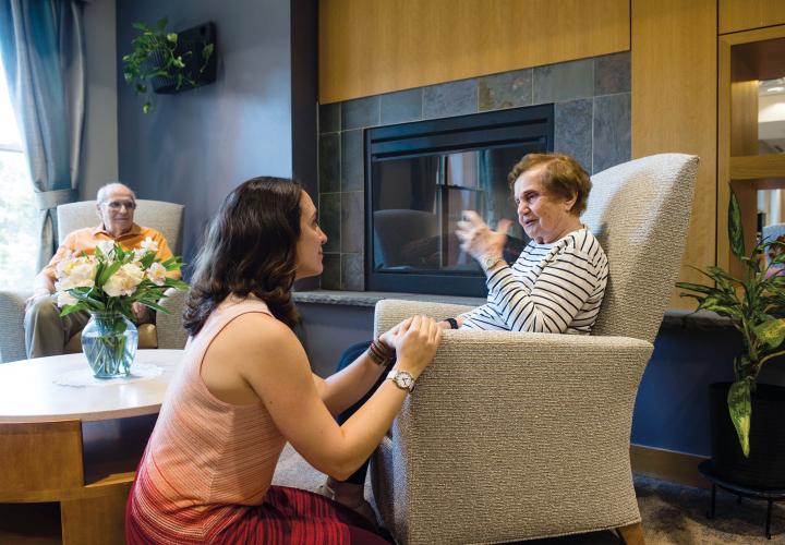 Younger woman kneels to listen to a seated older relative reminisce.