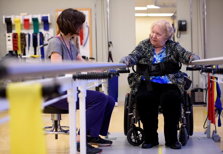 A patient in a wheelchair rehabs at bars with seated therapist.