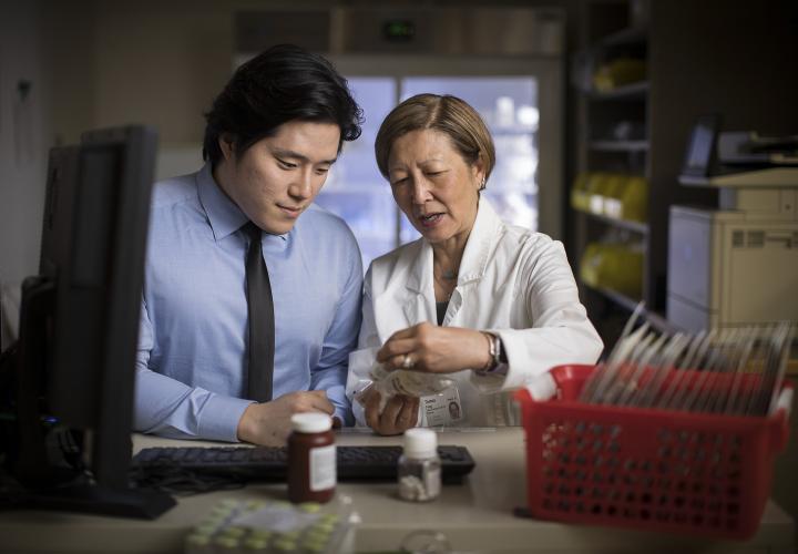 A female pharmacist in a white coat is standing next to a young man; they are in a pharmacy at Hebrew Rehabilitation Center, looking at a piece of paper.