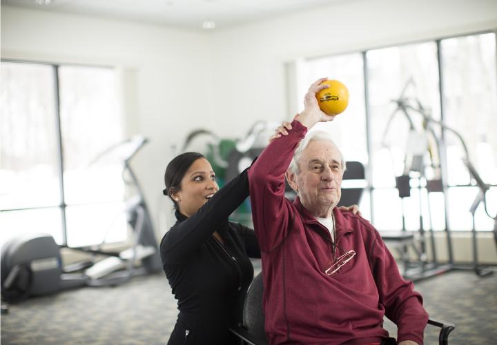 A senior holds a ball above his head with the help of a physical therapist in the Simon C. Fireman Community fitness center.