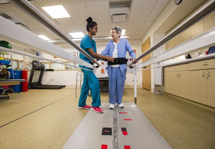 Physical therapist helps older woman walk along balance bars in rehabilitation gym at Hebrew Rehabilitation Center