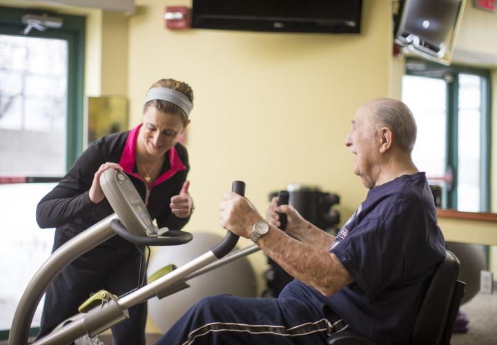 An older man is seated using a recumbent elliptical machine. A younger woman is standing next to him, looking at the display on the machine, smiling and giving him a thumbs up.