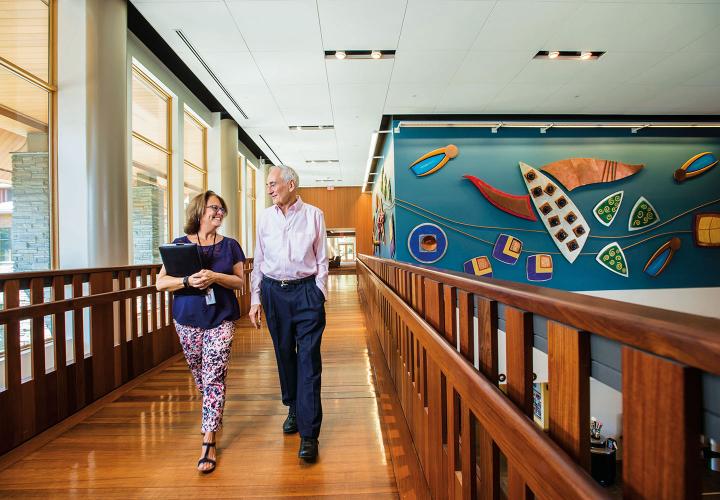 An older man in an oxford shirt and navy trousers walks across the community center bridge with the NewBridge on the Charles move-in coordinator.