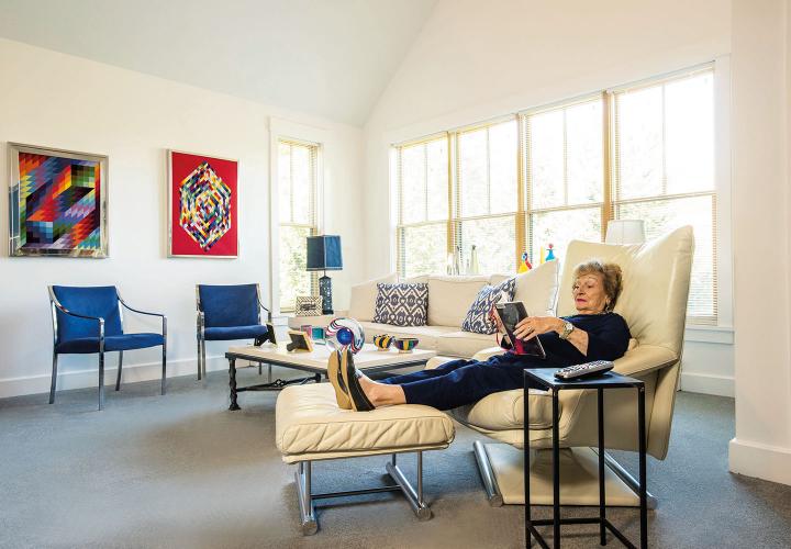 An independent living resident reads a book as she reclines on a white leather chair in her bright, contemporary cottage residence at NewBridge on the Charles.