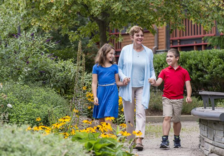 An older woman holding the hands of a younger boy and younger girl; they are walking through a flower garden at NewBridge on the Charles