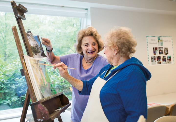 Two Orchard Cove residents stand at an easel, discussing a painting in progress, in a sunny art studio.