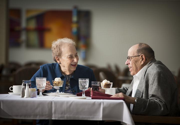 An older man and woman are seated at a restaurant table, smiling and looking at each other. There is dessert in front of them and the woman is holding a spoon.