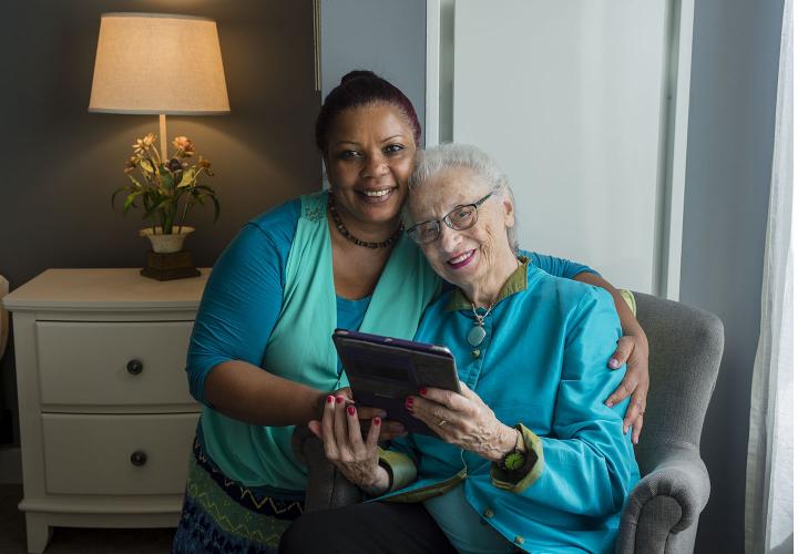 Smiling caregiver with arm around older adult woman who is smiling and holding an iPad