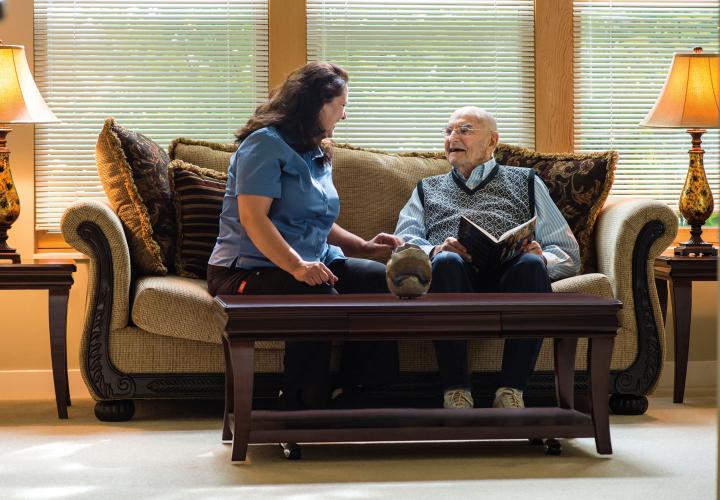An older man laughs with a nursing assistant in a well-furnished living room.