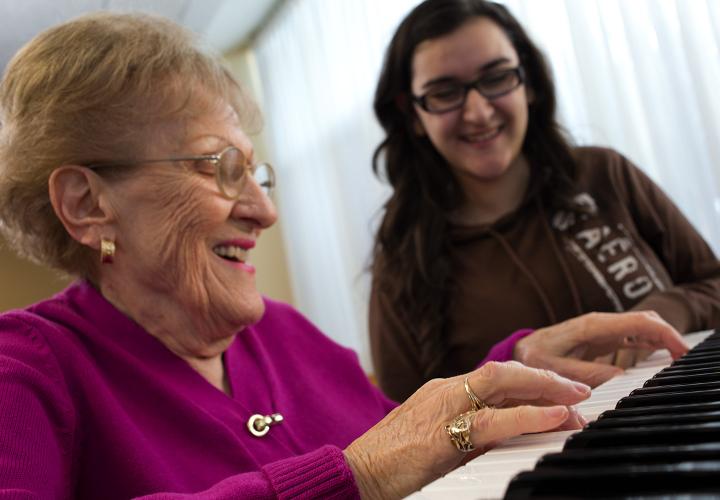 Older woman in pink sweater plays a duet on piano with a high school student