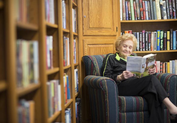 A woman sits in an easy chair, smiling, reading a book, surrounded by walls of bookshelves.