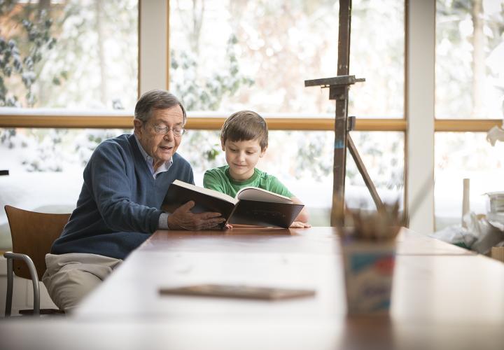 Older man in blue sweater and grade-school student in green t-shirt read through a large book together.