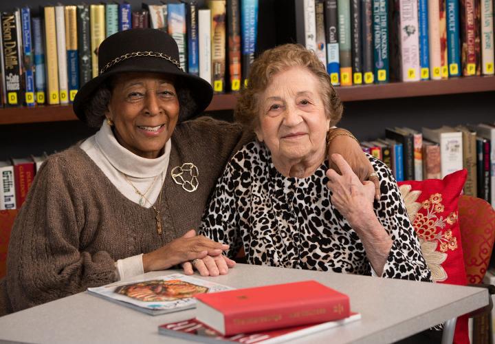 Two older women sit together in a library at a table with books and magazines.