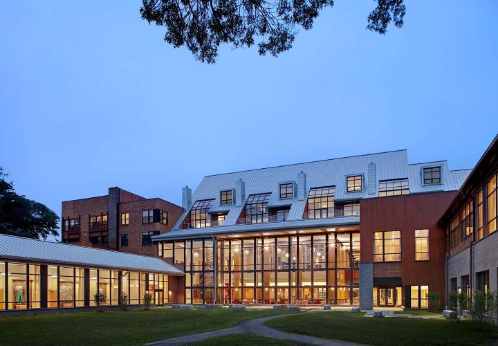 An exterior view of the Shapiro Community Center at NewBridge on the Charles at dusk, with multi-story windows revealing contemporary décor within.