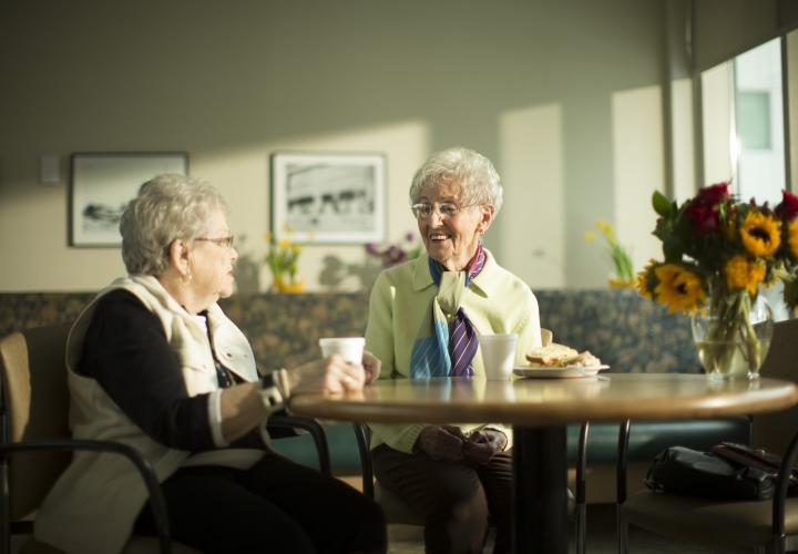 Two women sit at a round table with styrofoam cups in front of them. One has a sandwich. They are smiling and talking. There is a vase of flowers in the foreground and a large window on their right.