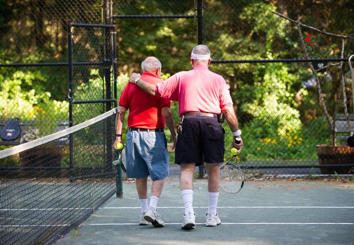 Two older men in colorful T-shirts share a moment as they walk off the tennis court after a game on the Orchard Cove campus.