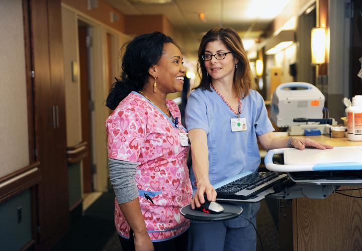 Two nurses stand in a hospital-like hallway, looking at a computer screen.