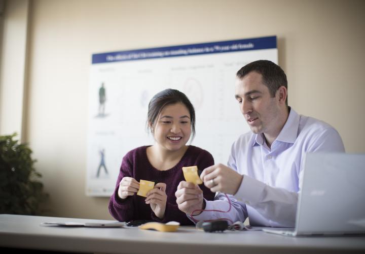 A female and a male researcher from the Hinda and Arthur Marcus Institute for Aging Research sit at a table with a laptop. They are each holding a small yellow square that has wires attached.
