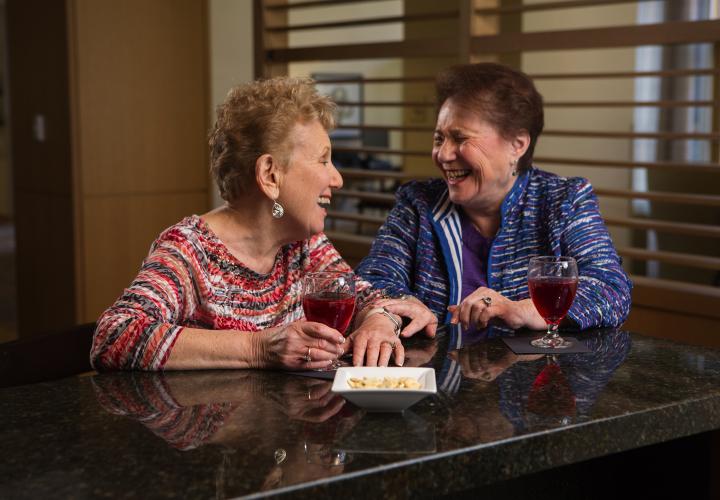 Two older women standing at a counter with juice in front of them; they are looking at each other and laughing.