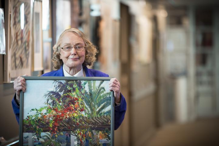 Older woman in a blue jacket proudly displays her painting.