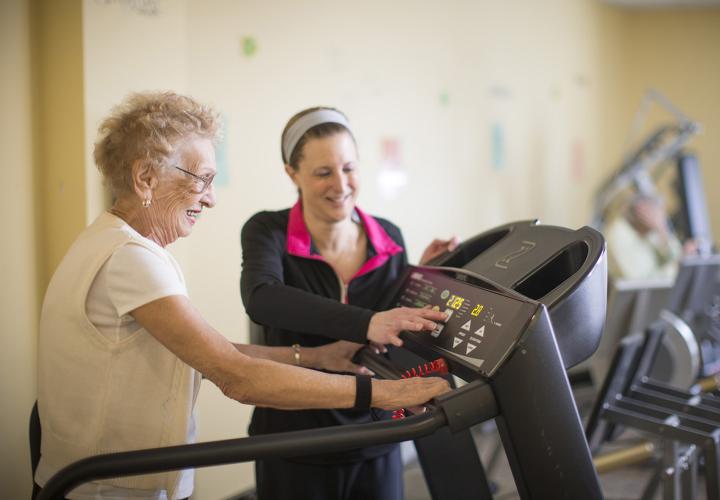 An older woman is walking on a treadmill. A younger woman in workout clothes stands next to her, changing the display on the treadmill. They are both smiling.