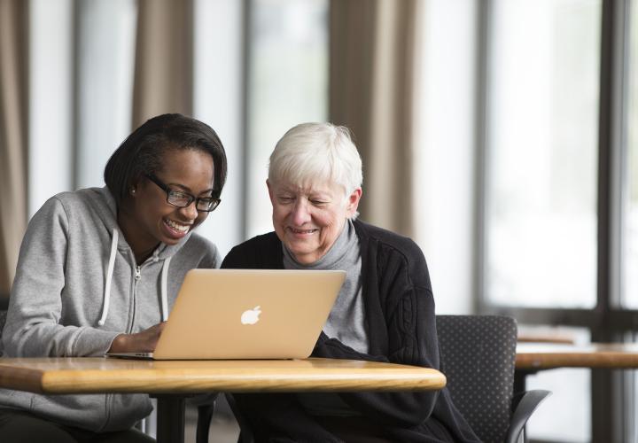 And older woman and a younger woman sit side by side at a table, smiling and looking at a laptop screen.