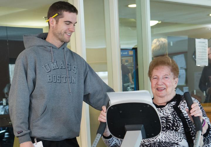 Hebrew SeniorLife fitness trainer helps an older woman exercise on a machine designed for geriatric fitness.