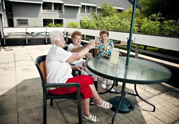 Three older women sit outdoors at a patio table, smiling and toasting with lemonade.