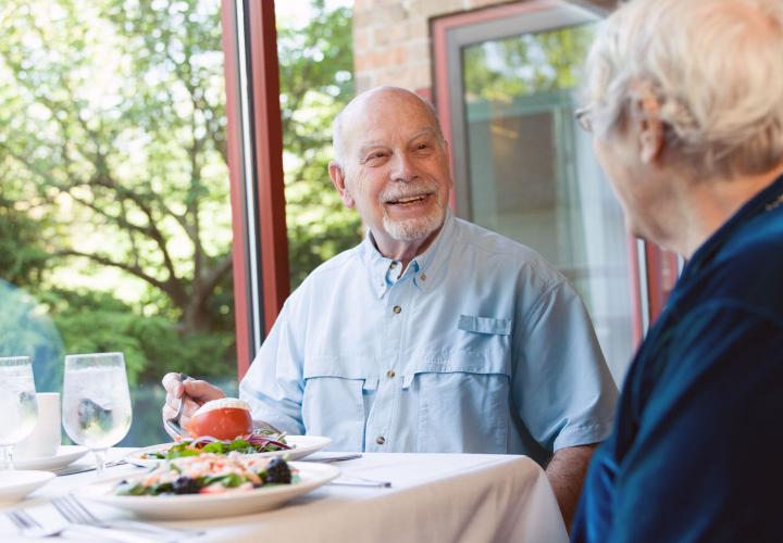 A older man in a blue shirt and a woman in a blue shirt share a meal in Orchard Cove’s Garden Room restaurant.