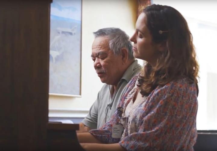 An older man sits at a piano next to a woman, playing the piano together as a part of music therapy.