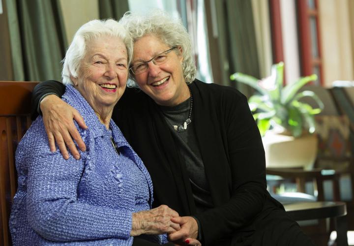 An adult daughter with her arm around and her mother's shoulder; both are smiling.