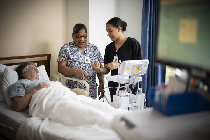 A registered nurse explains the automatic blood pressure machine to a certified nurse assistant as a patient looks on.